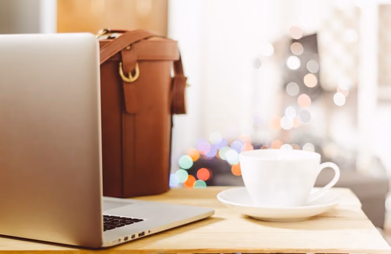 Laptop beside a brown leather bag and a white teacup on a wooden table with blurred lights background.