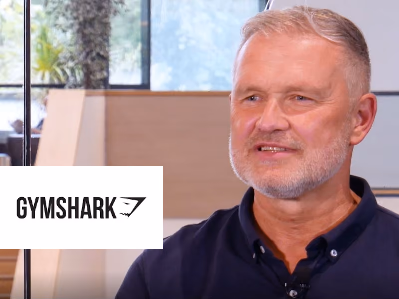 Middle-aged man with grey hair and beard speaking indoors near a glass window and Gymshark logo.