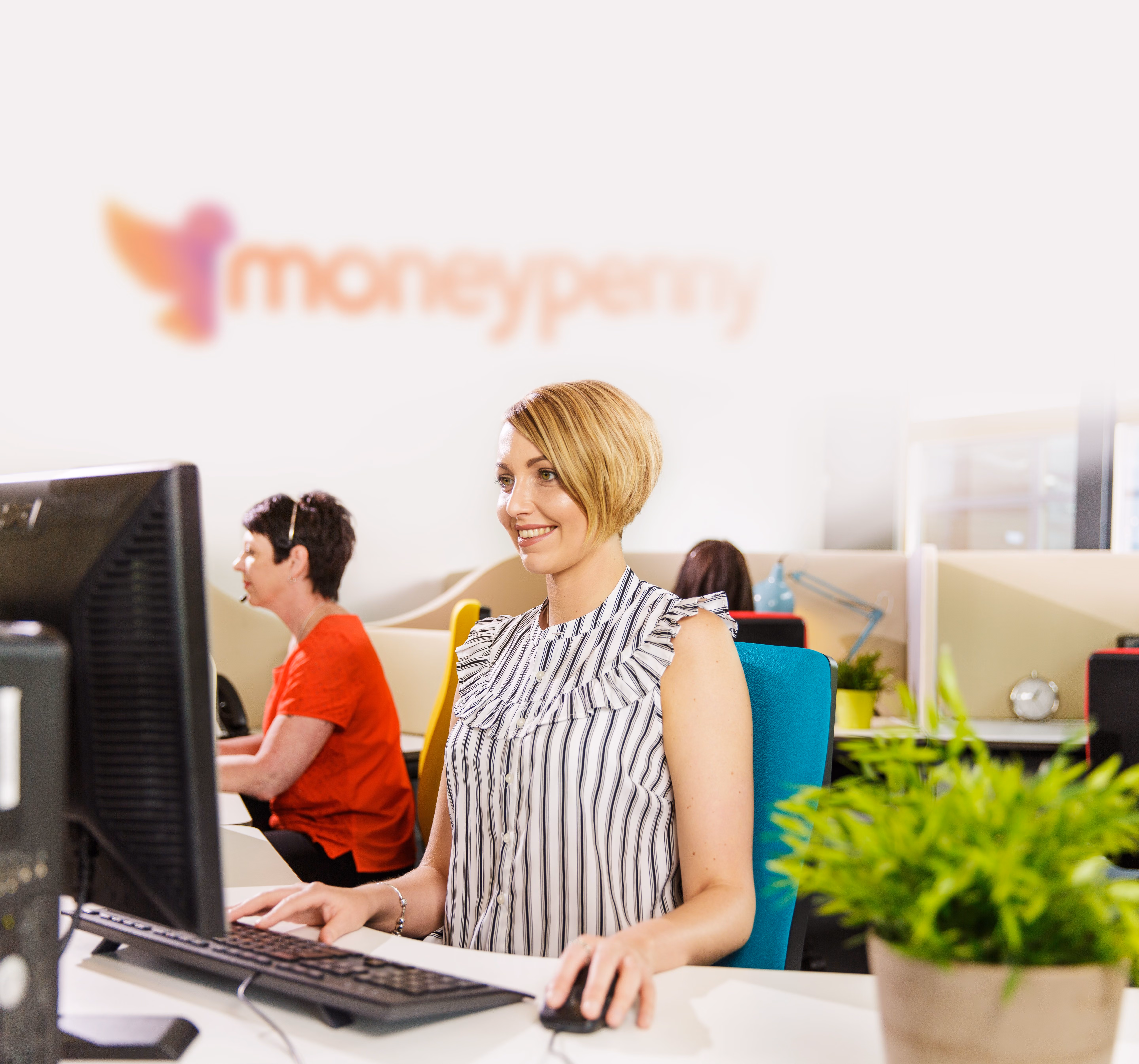 Smiling woman with short blonde hair working on a computer in an office with other employees.