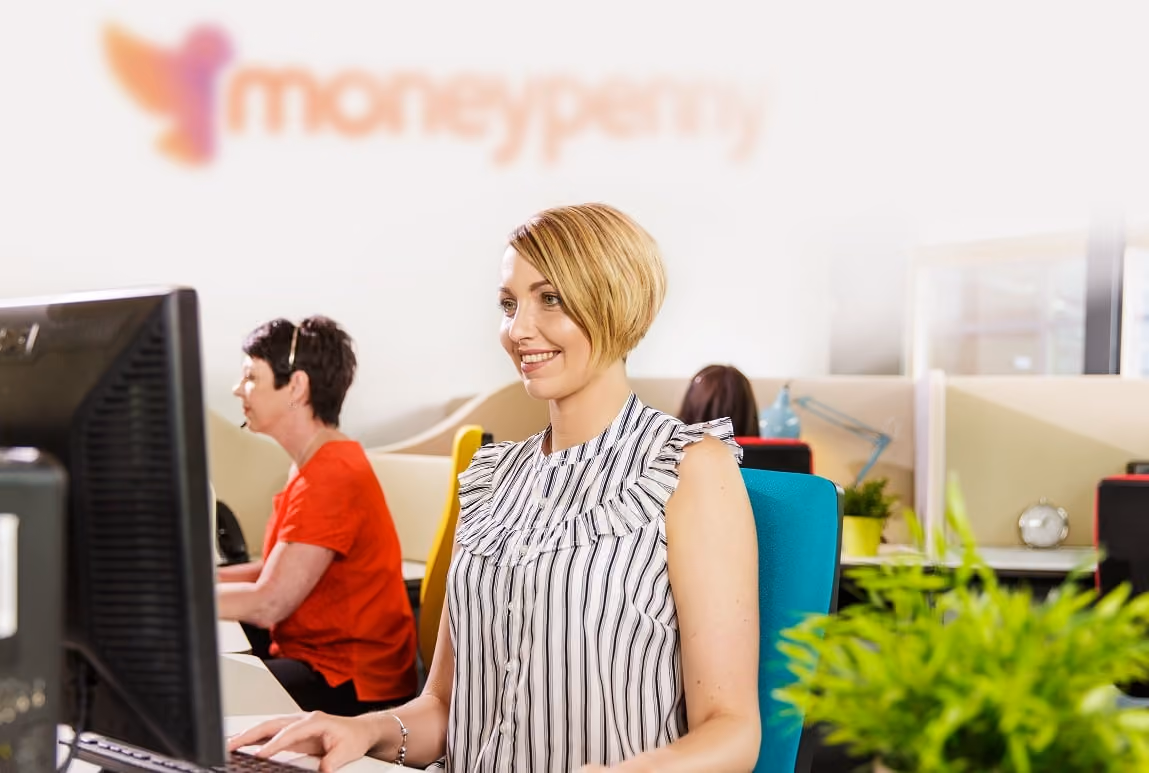 Smiling woman with short hair working on a computer in a bright office with colleagues behind her.