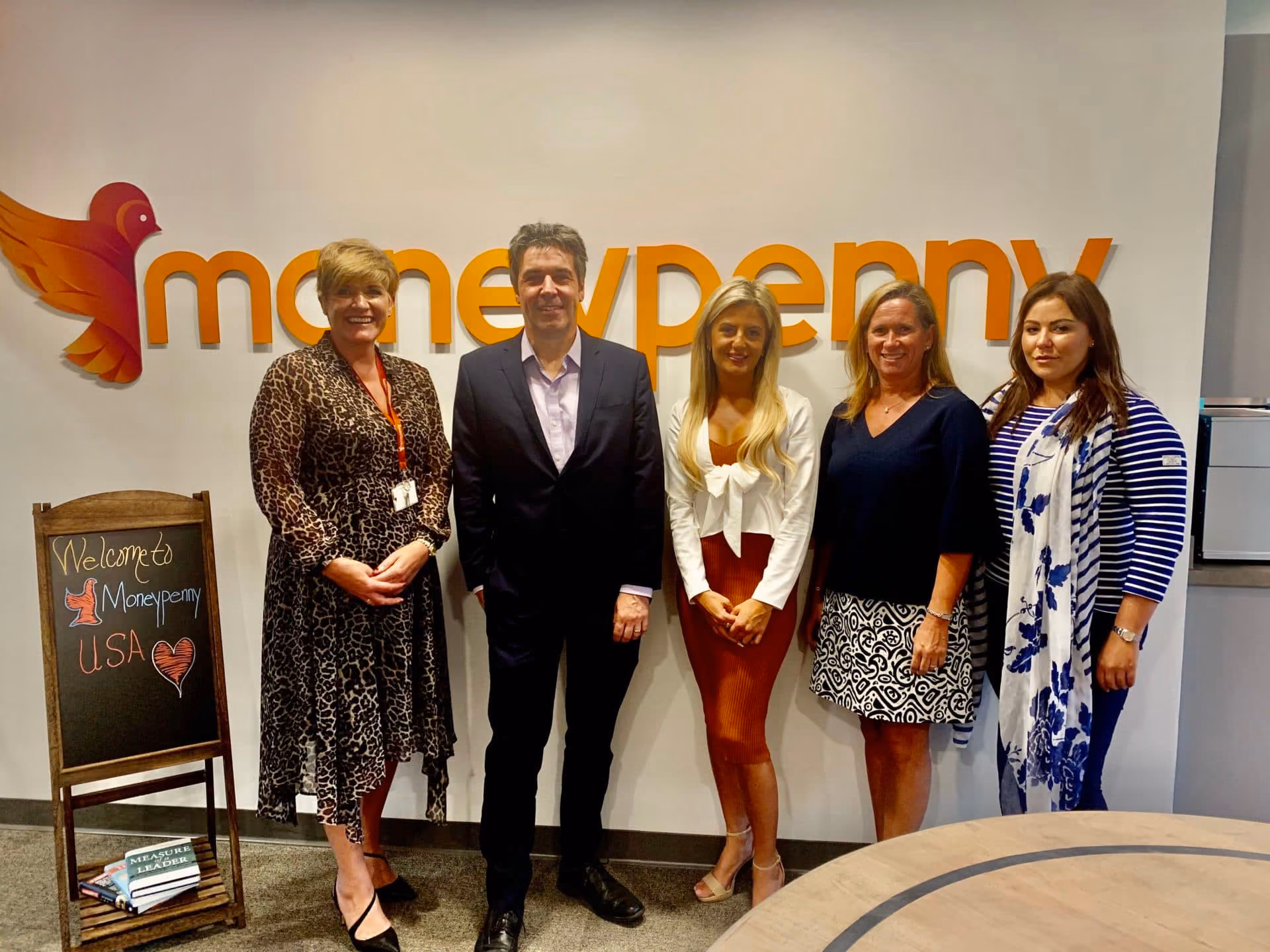Five people standing in front of Moneypenny logo with welcome sign and books nearby.