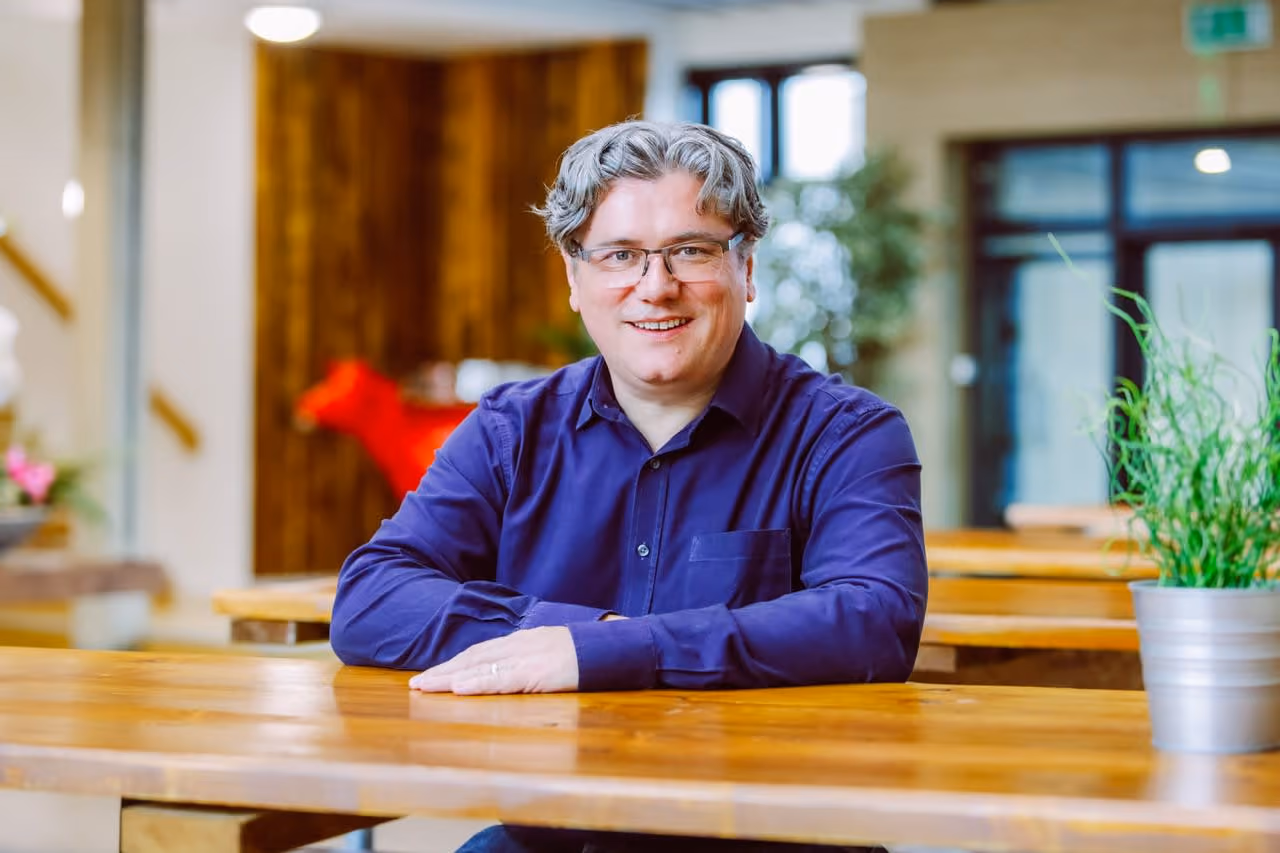 Man with glasses and gray hair sitting at a wooden table smiling in a bright room with plants.