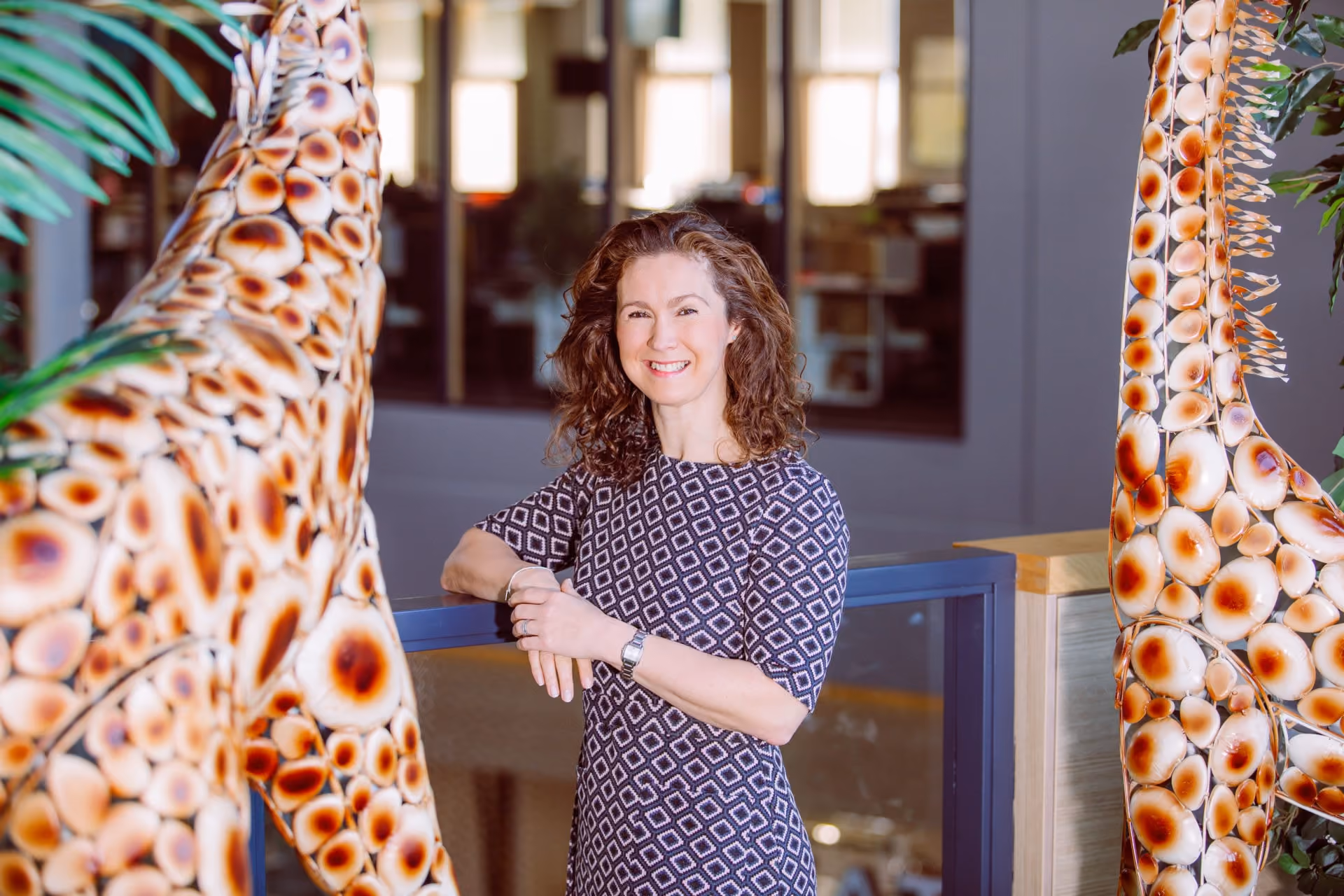 Smiling woman with curly hair in a patterned dress leaning on a railing indoors between giraffe sculptures.