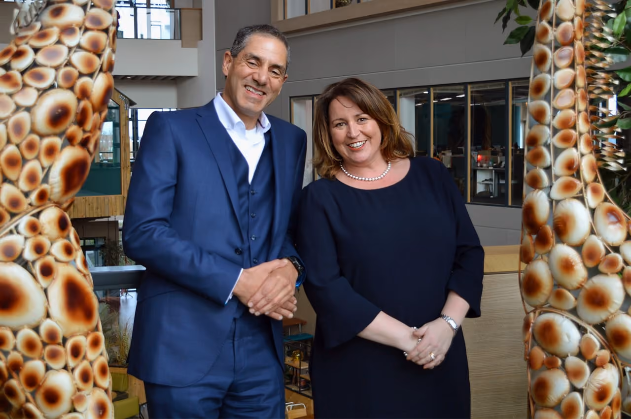 Man in blue suit and woman in black dress smiling side by side in modern office setting.
