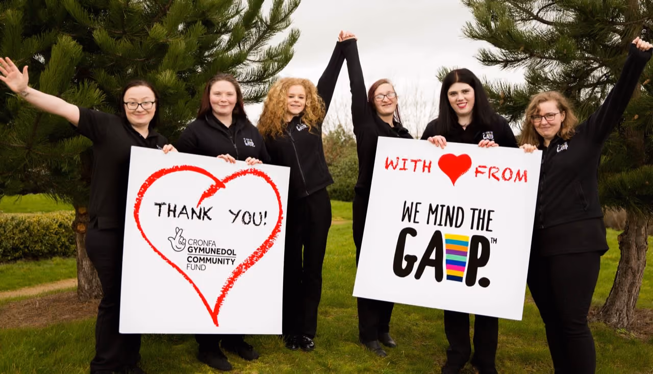 Six people outdoors holding signs: one with 'Thank You' heart, the other says 'With Love From We Mind The Gap'.