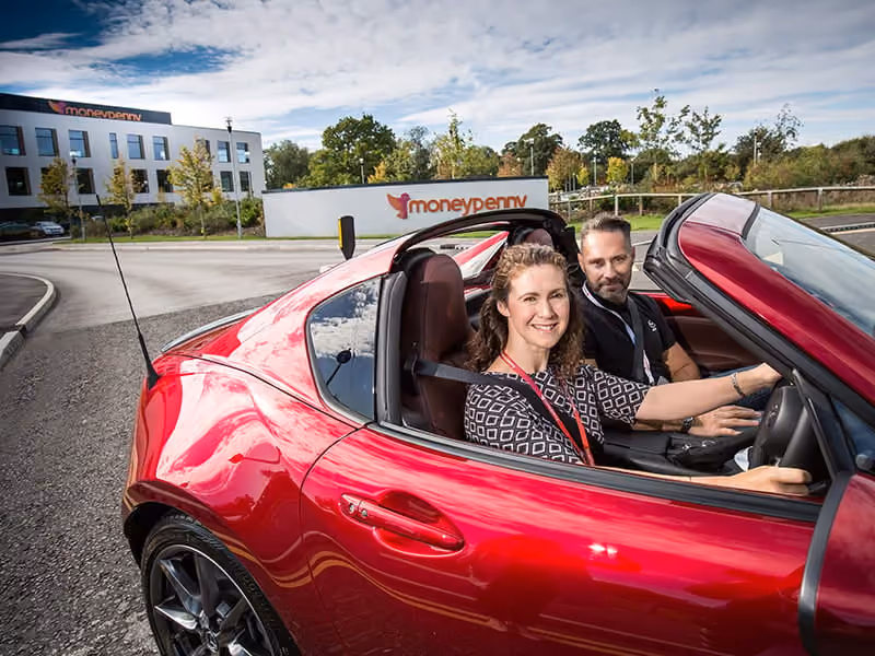 Smiling woman driving a red convertible car with a man seated beside her outside Moneypenny office.