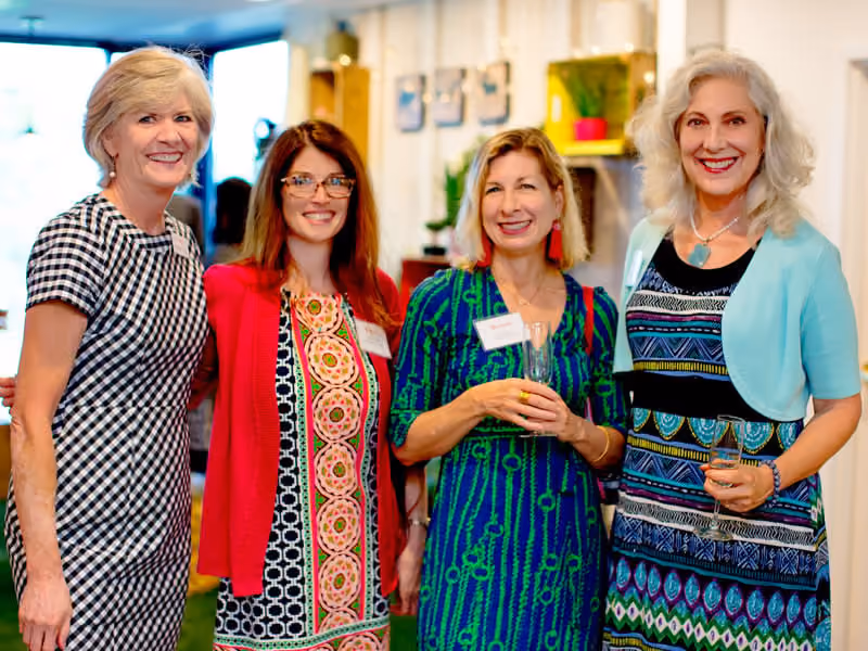 Four smiling women wearing colorful dresses and holding drinks in a bright indoor setting.