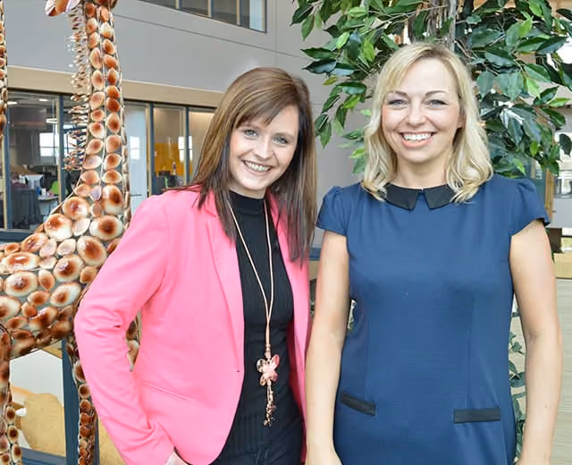 Two smiling women standing indoors near a large giraffe sculpture and green plant.