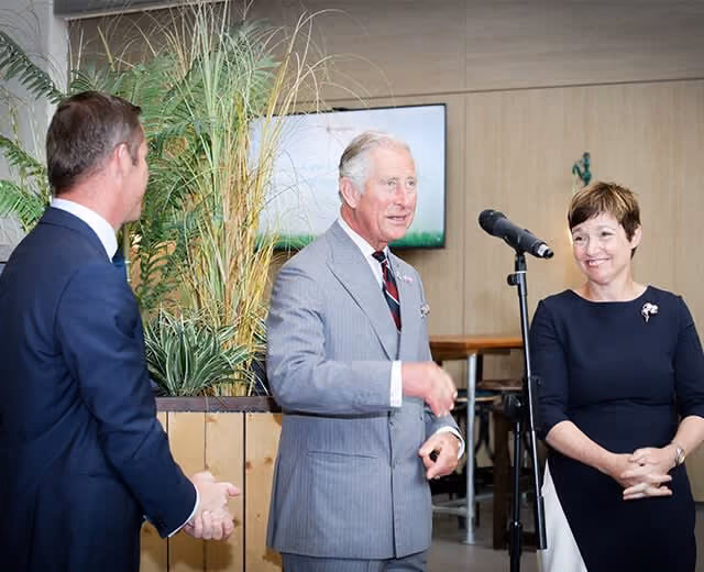 Three people in a formal setting; man in gray suit speaking, woman smiling, another man listening.