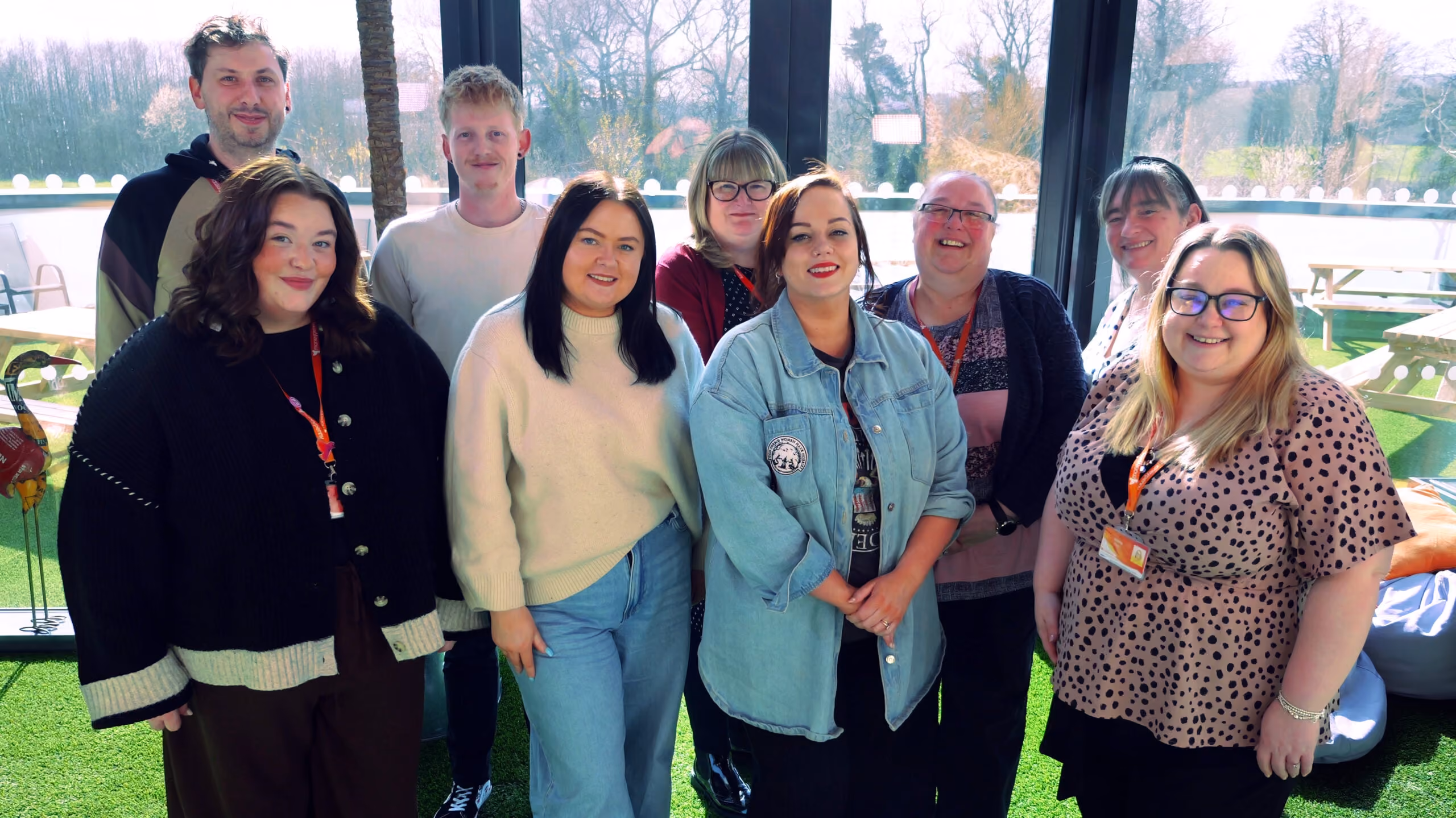 Group of nine adults standing closely indoors with large windows and outdoor view behind them.