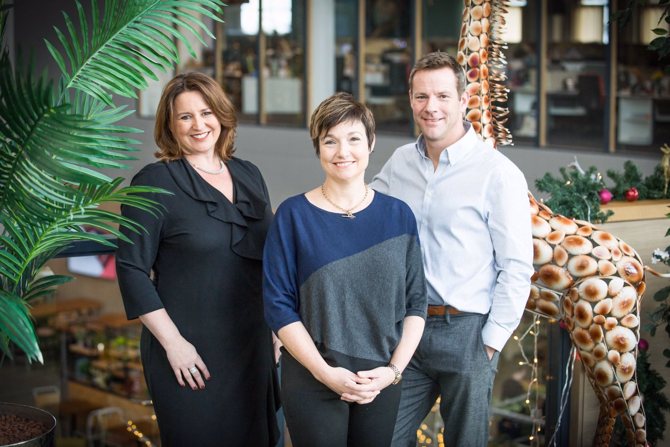 Three adults smiling indoors with plants and a decorative giraffe sculpture in the background.