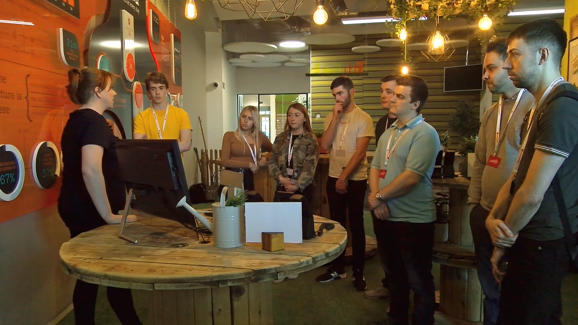 Woman presents to a group of attentive young adults standing around a rustic round wooden table with a computer.