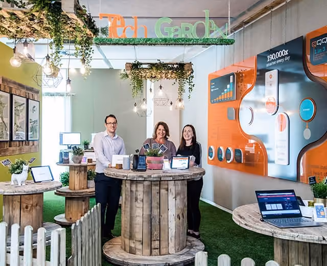 Three people standing behind wooden spool tables with tech displays in a green-themed indoor tech garden.