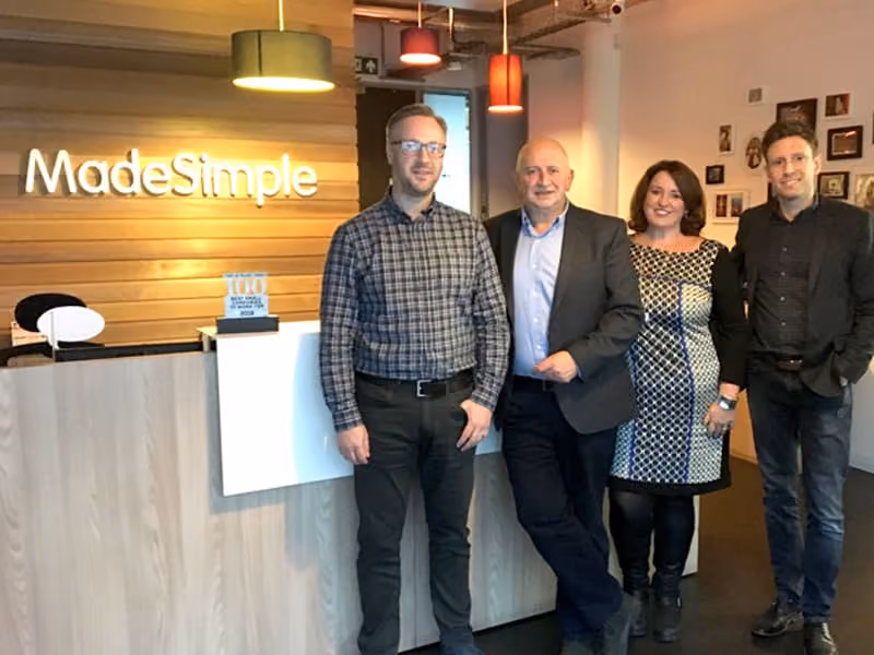 Four people standing and smiling in an office lobby with a MadeSimple sign on a wood-paneled wall.