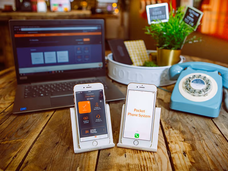 Two smartphones on stands displaying Pocket Phone System app in front of a laptop and a vintage blue rotary phone.