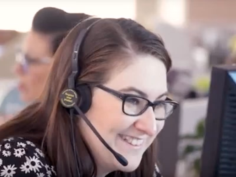 Smiling woman with glasses wearing a headset, talking at her computer in an office.