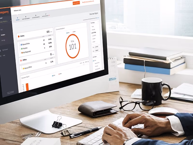 Hands typing on keyboard at desk with glasses, coffee cup, wallet, and a screen showing a dashboard with data.