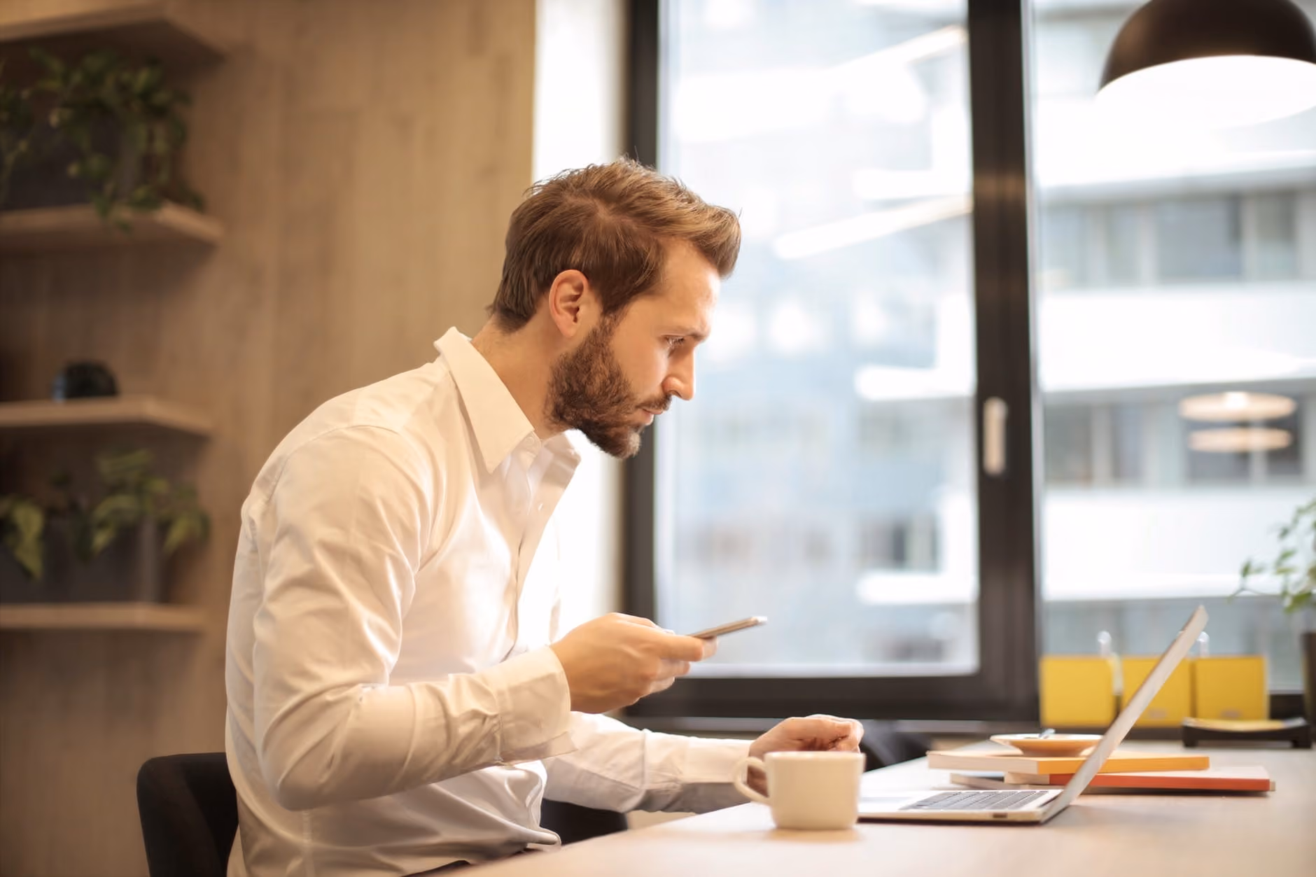 Man in white shirt seated at desk using smartphone with laptop and coffee cup nearby in office.