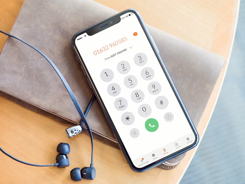 Smartphone showing dial pad with phone number 01632 960585 on a table next to black earbuds.
