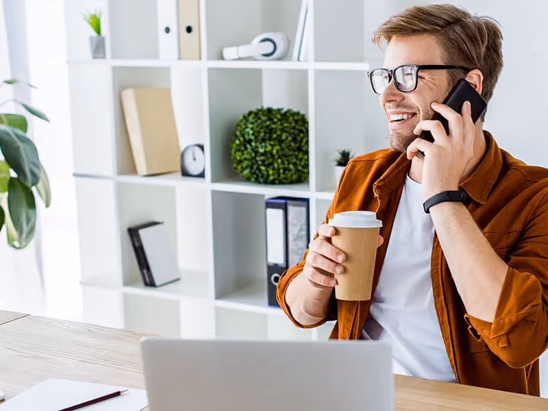 Smiling man wearing glasses holding coffee cup, talking on phone in bright modern office.