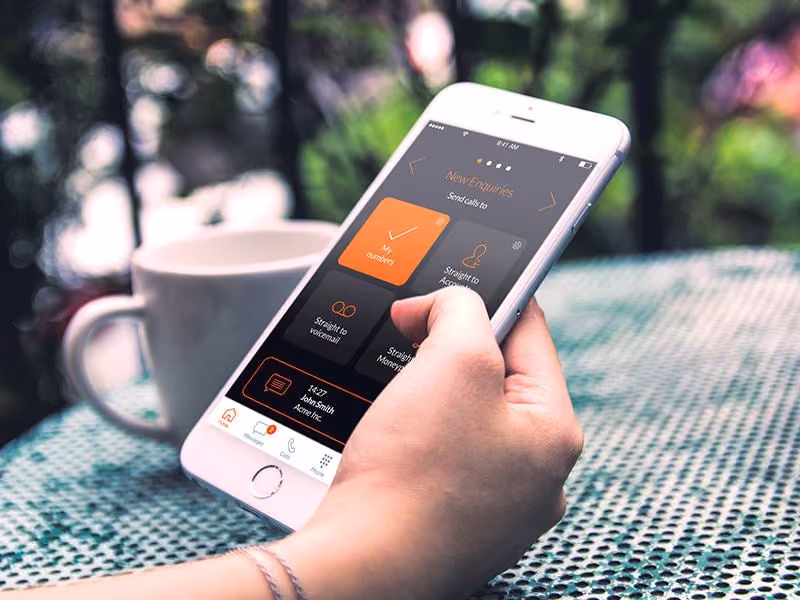 Hand holding smartphone showing app screen with call options, table and coffee cup in background.