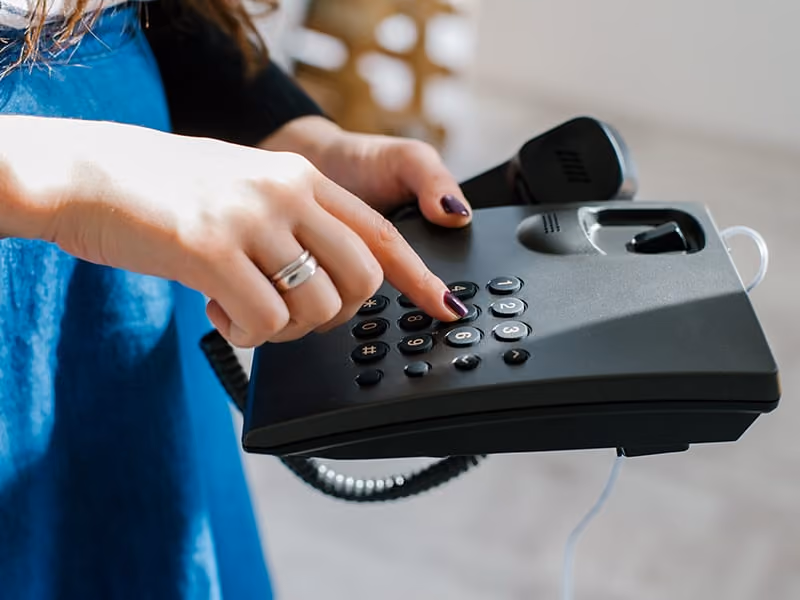 Person with dark nail polish dialing a number on a black office telephone keypad.