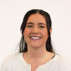Smiling young woman with long dark hair wearing a white top against a plain light background.