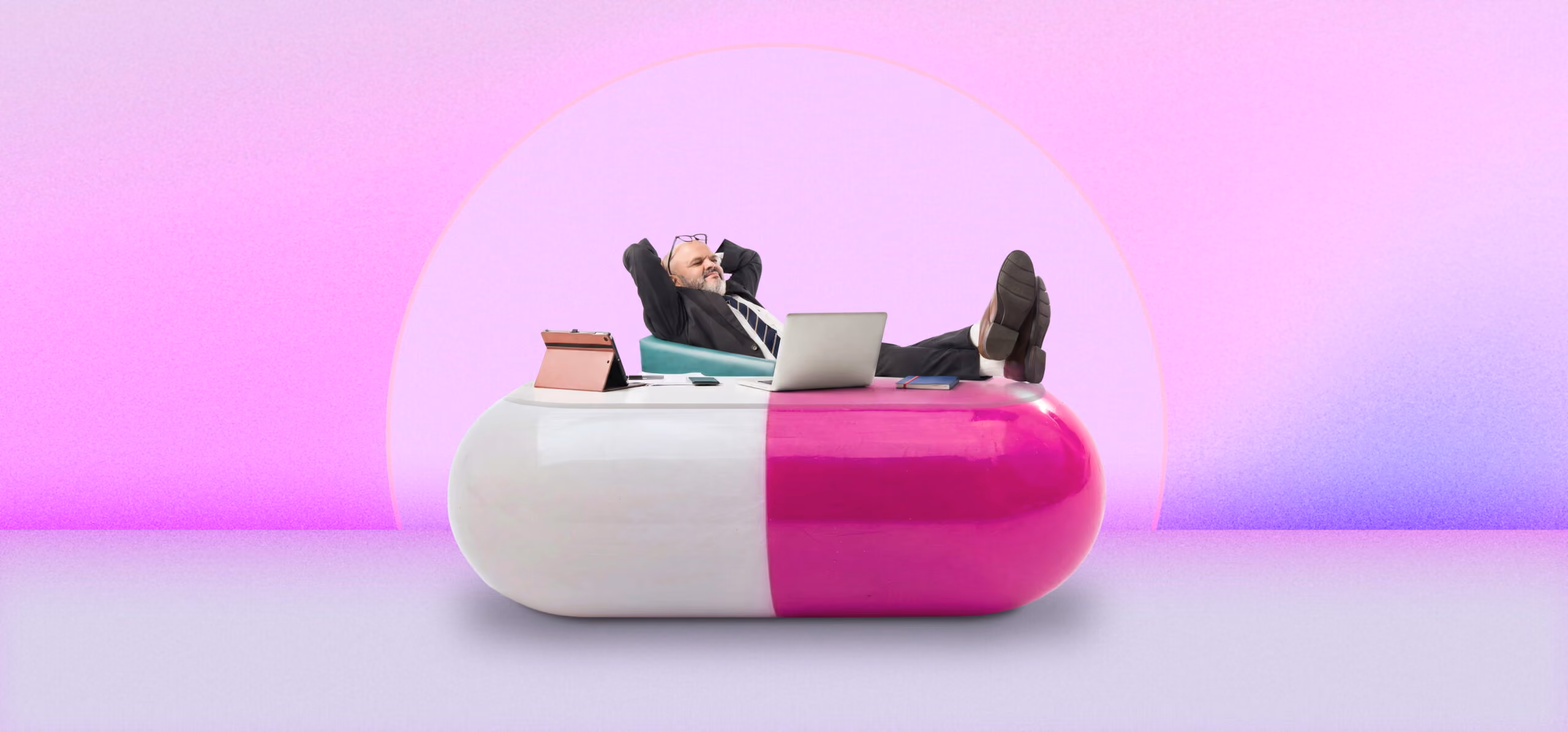 Man in suit reclining with feet up on a giant pink and white capsule-shaped desk with laptop.