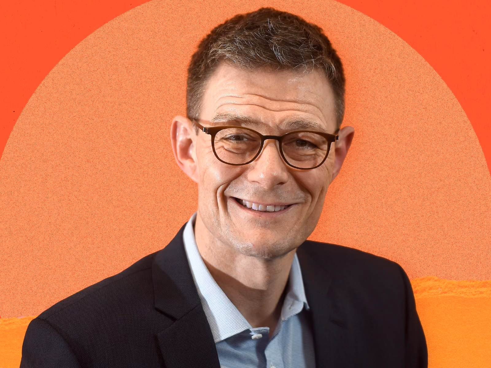 Smiling man wearing glasses and a dark blazer with a light blue shirt against an orange background.