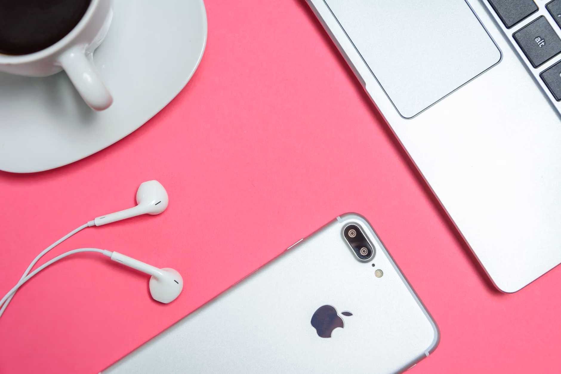Silver iPhone, white earphones, silver laptop keyboard, and coffee cup on pink surface.