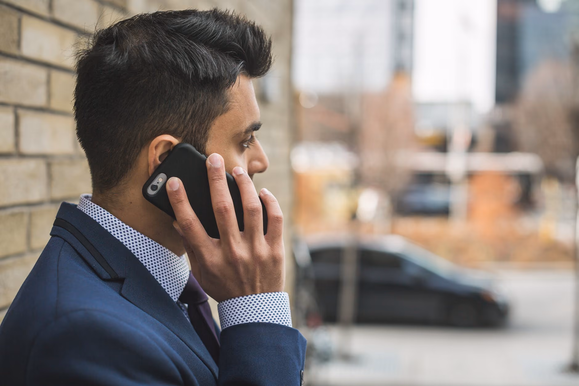 Young man in a blue suit talking on a smartphone outside near a brick wall in an urban setting.