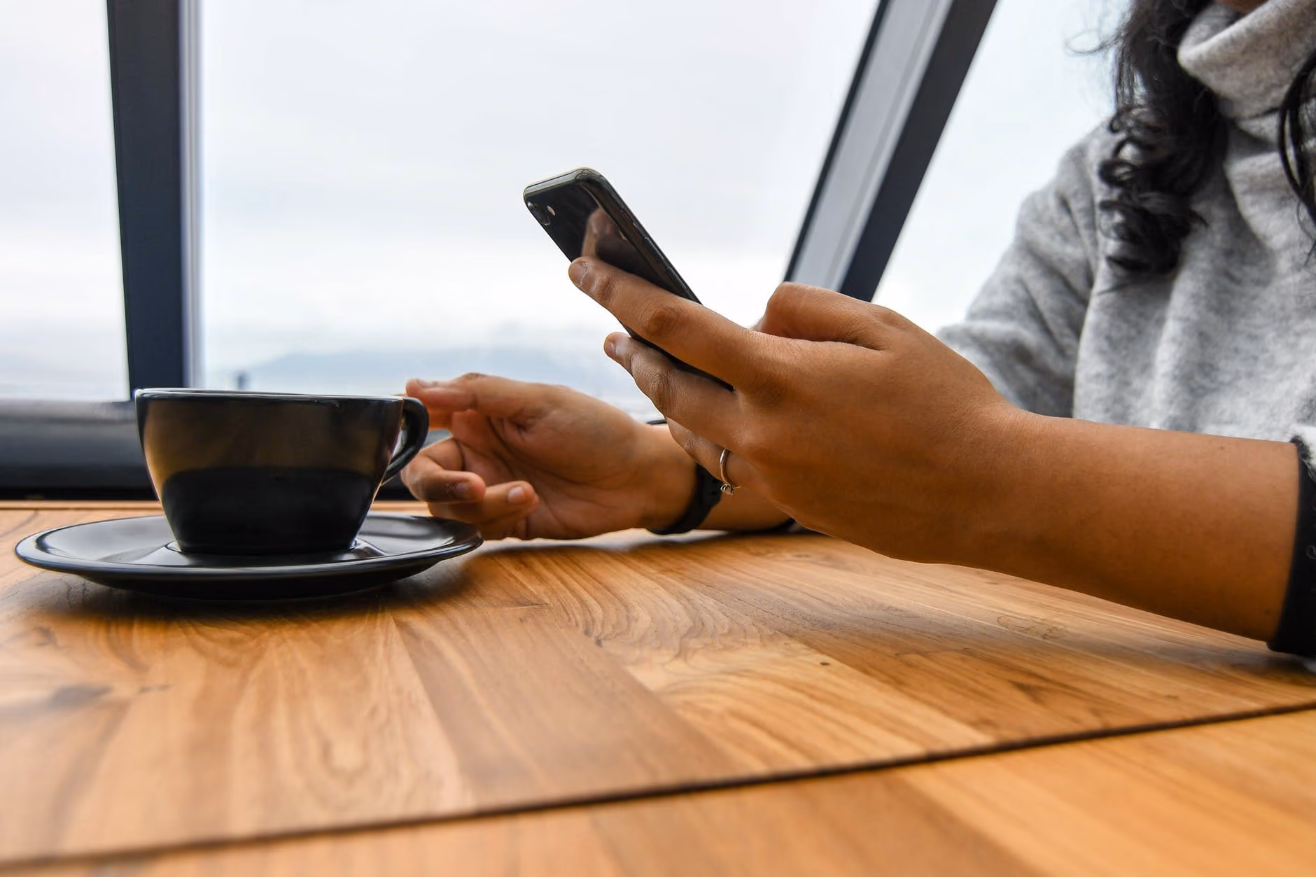Person holding a smartphone with one hand and reaching for a black coffee cup on a wooden table.