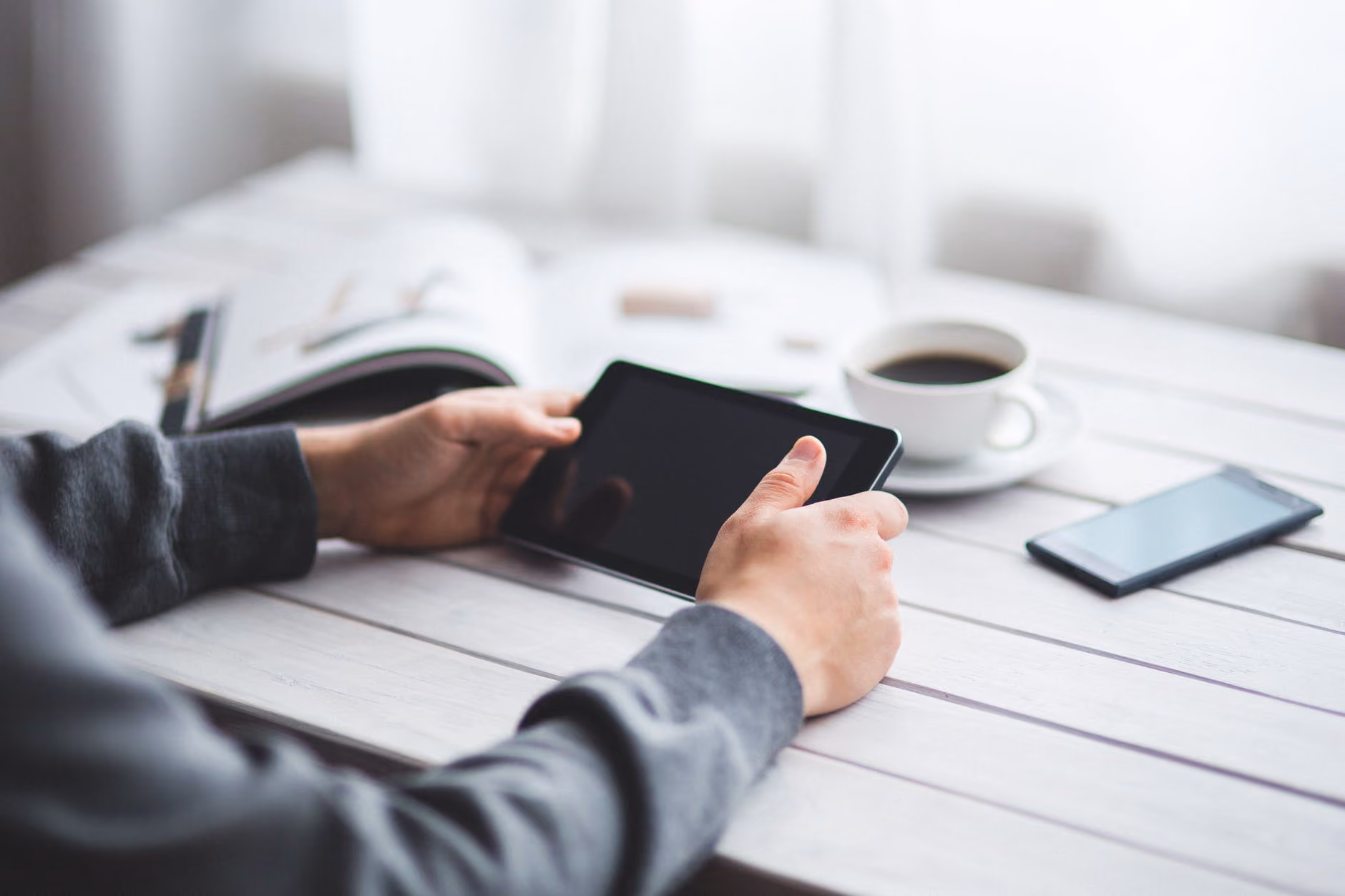 Person holding a black tablet on a white wooden table with a smartphone, coffee cup, and open book nearby.