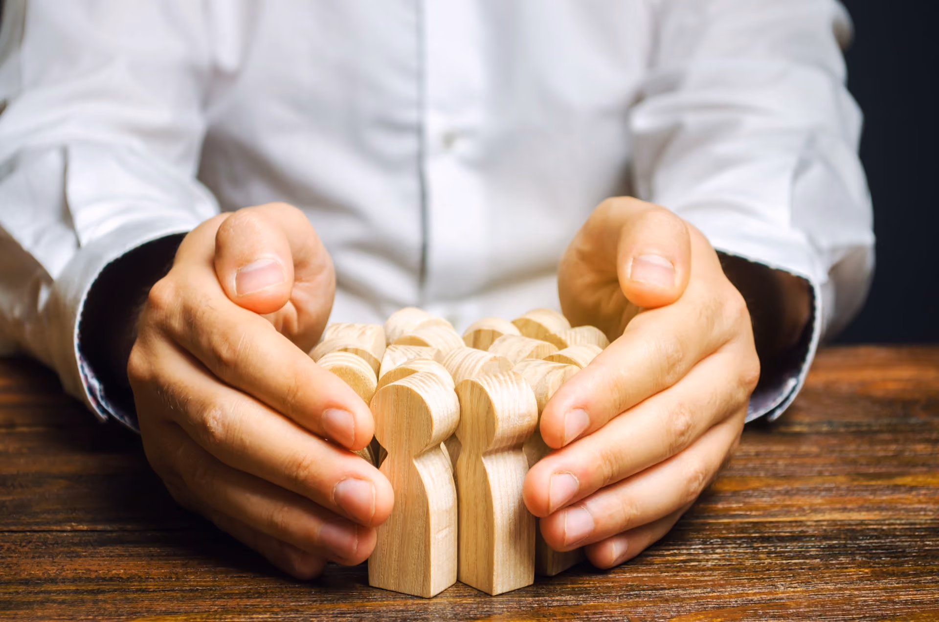 Hands in white shirt protect a group of wooden human figures on wooden table.