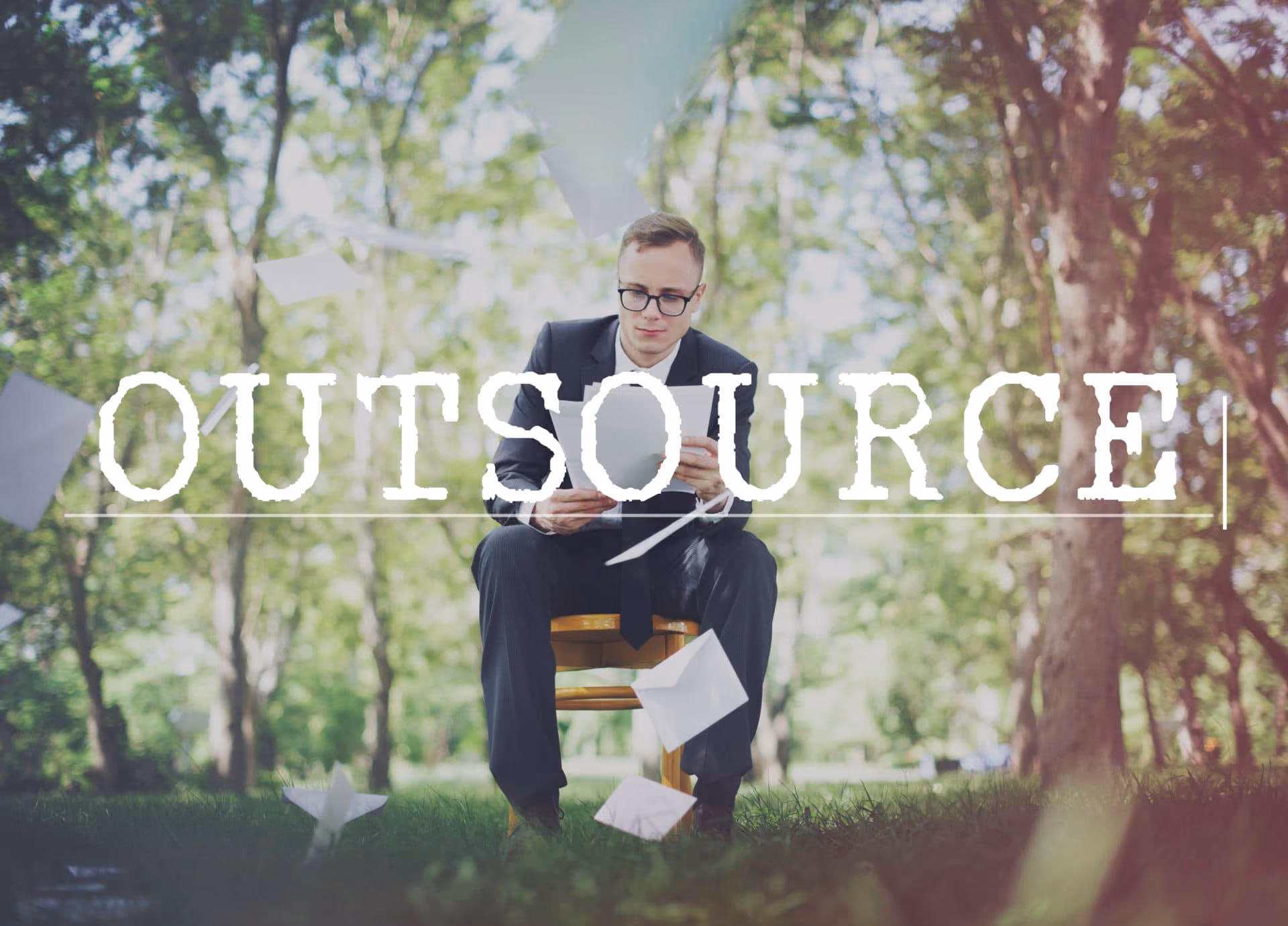 Man in suit sitting on chair outdoors reading papers as sheets fly around with word OUTSOURCE overlay.