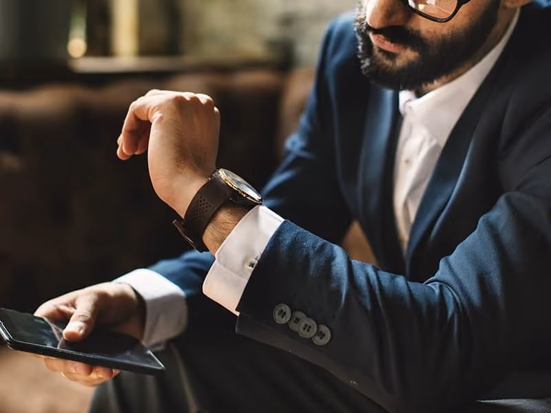 Bearded man in glasses and suit checking his wristwatch while holding a smartphone.