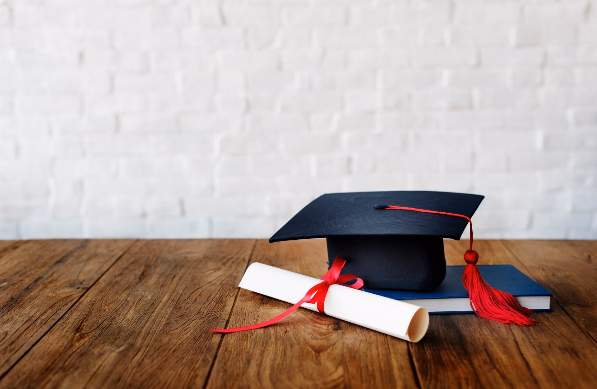 Graduation cap with red tassel on a blue book next to a rolled diploma tied with red ribbon on wood.
