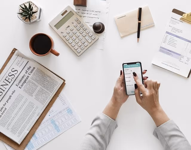 Hands holding smartphone displaying pay invoice screen on a desk with coffee, calculator, and documents.