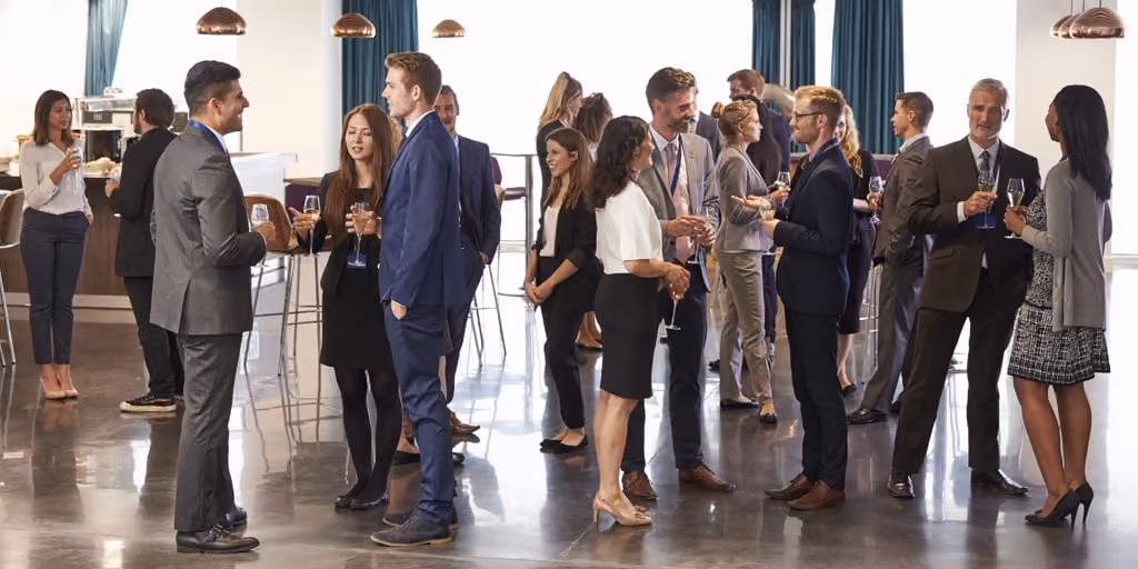 Business professionals in formal attire networking and chatting while holding drinks in a modern indoor space.
