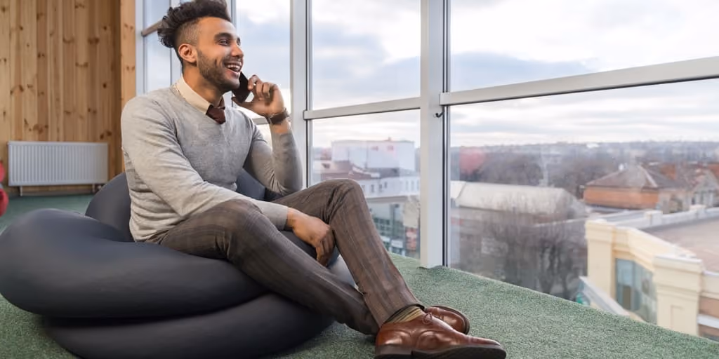 Man sitting on a black beanbag by a large window, smiling and talking on his phone.