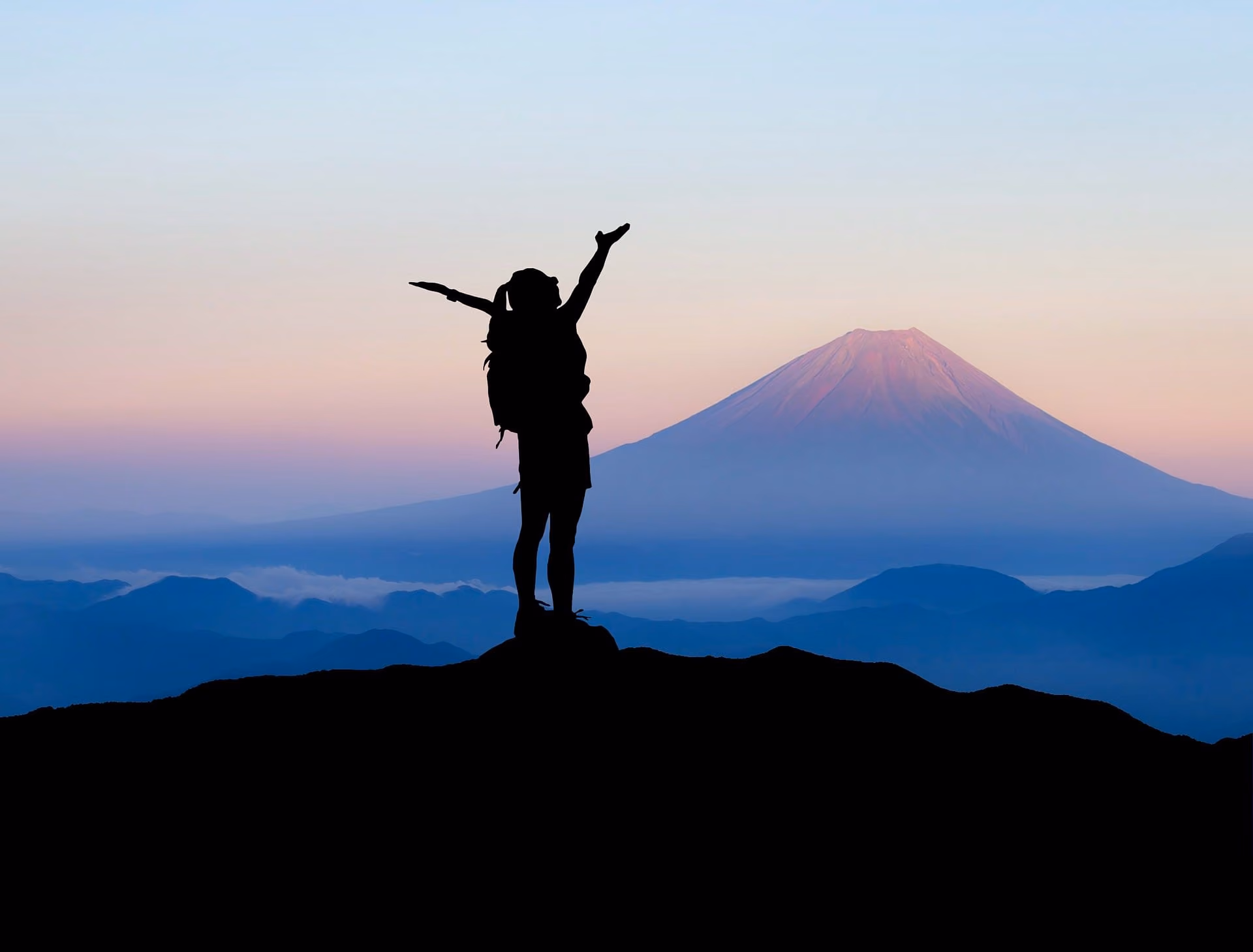 Silhouette of a person with a backpack raising arms on a hill at sunset with a mountain in the distance.