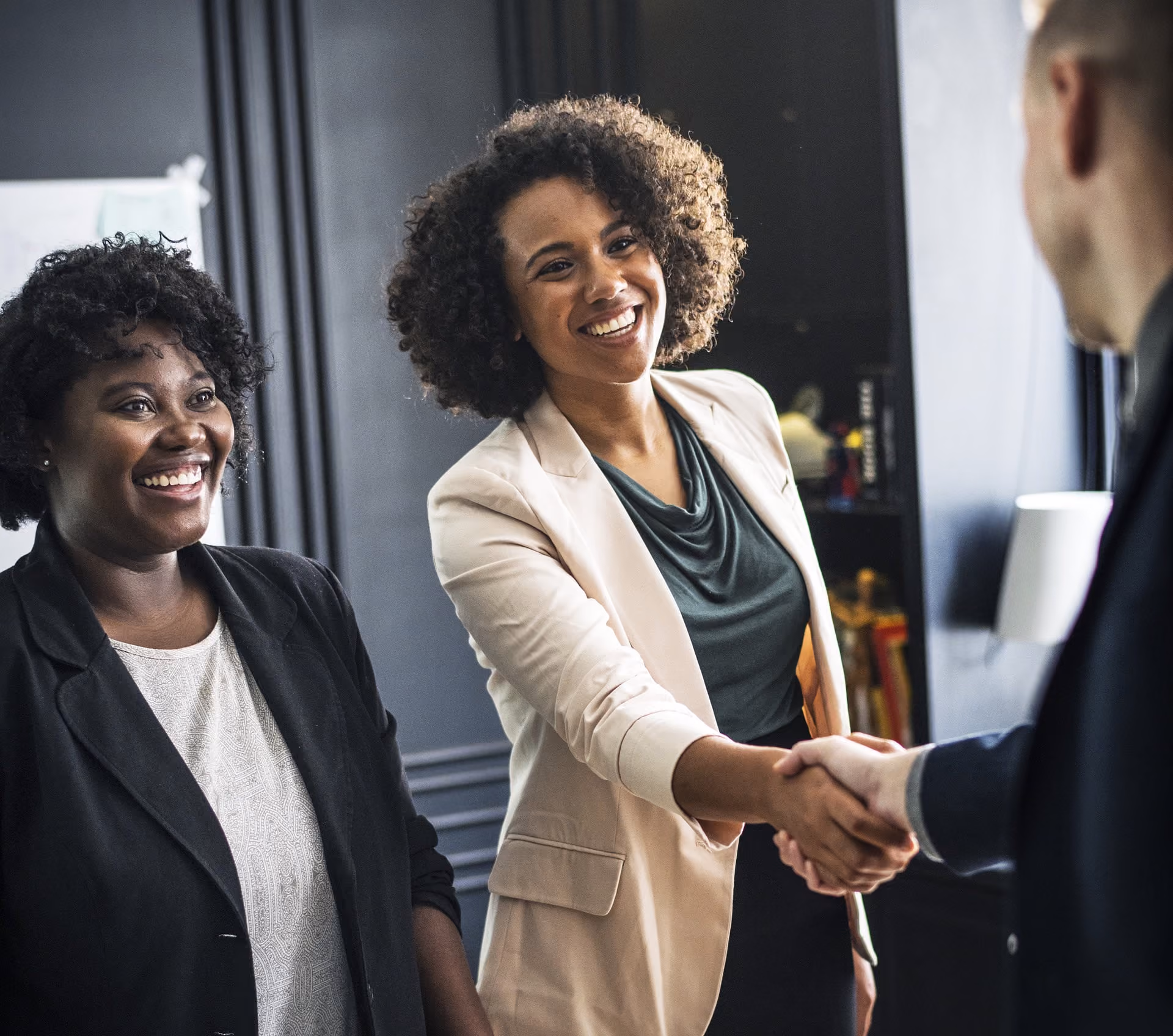 Two professional women smiling, one shaking hands with a man in an office setting.