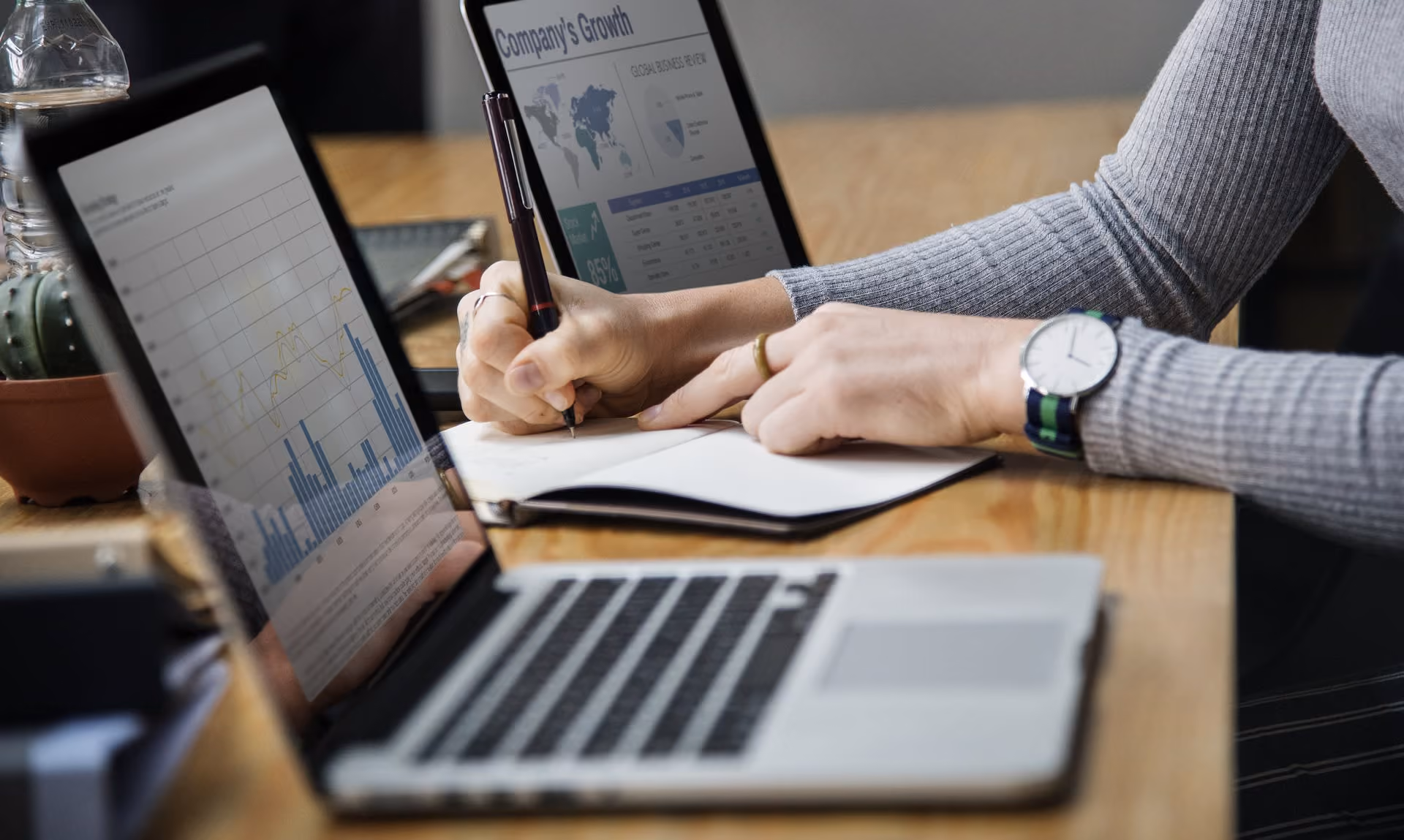 Person writing notes beside a laptop showing business growth charts and data on screen.