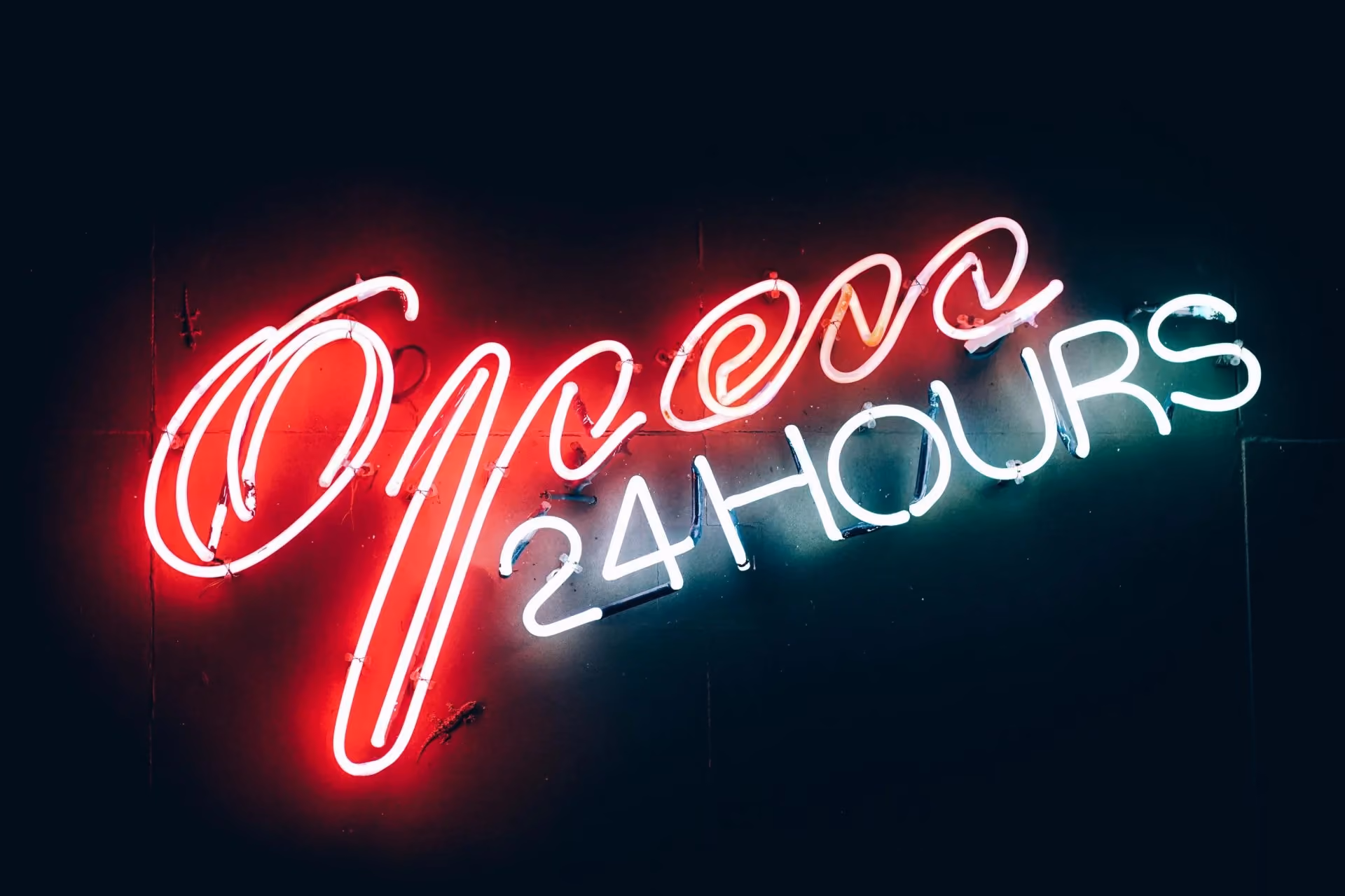 Neon sign glowing red and white reading "Open 24 Hours" on a dark background.