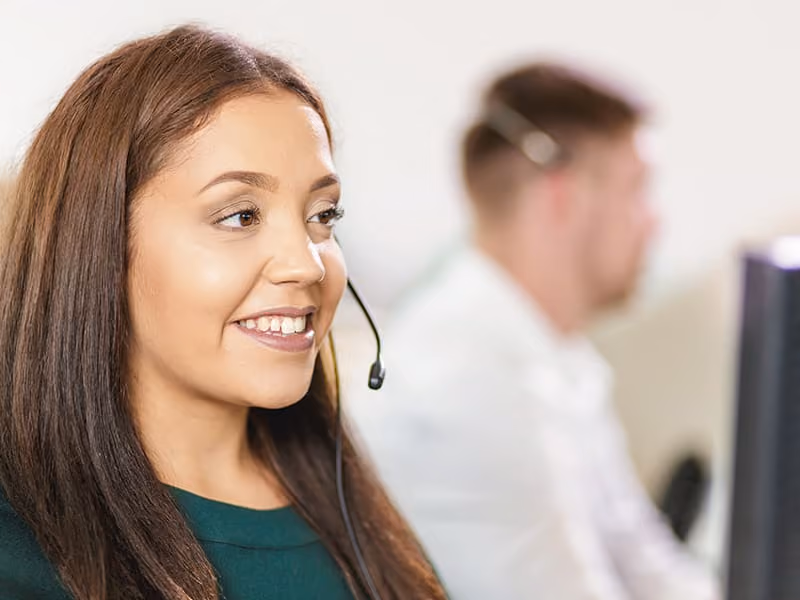 Smiling woman with headset working in a call center with a male coworker in the background.