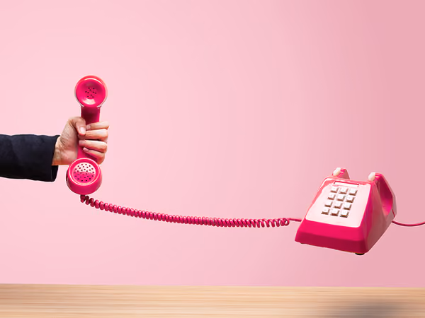 Hand holding the receiver of a pink vintage telephone with the phone base on a wooden surface.