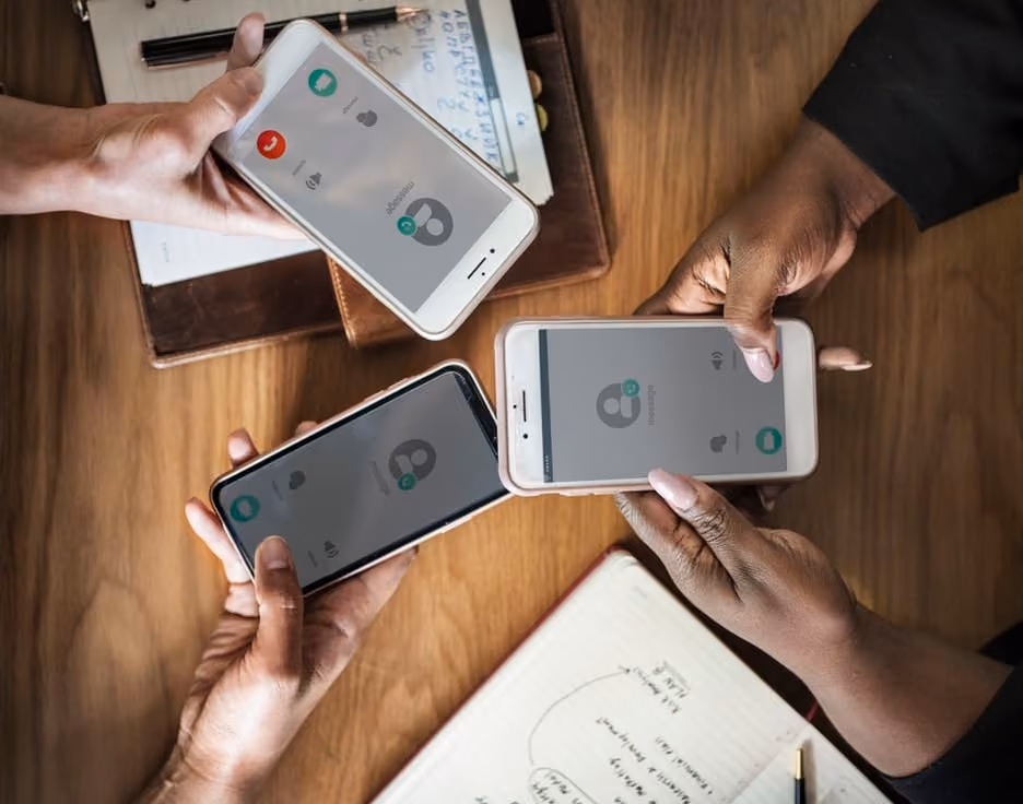 Three people holding smartphones displaying incoming calls, over a wooden table with notebooks and a pen.