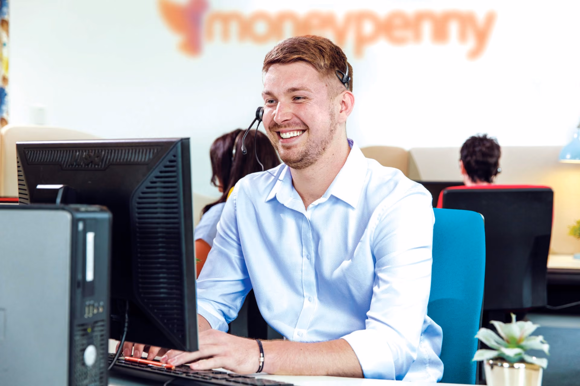 Smiling man with headset typing on keyboard in a bright office with others working in the background.
