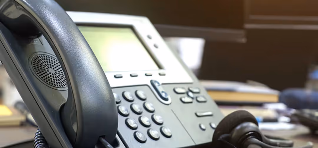 Close-up of a black office telephone with keypad, handset, and headset on a desk with blurred background.