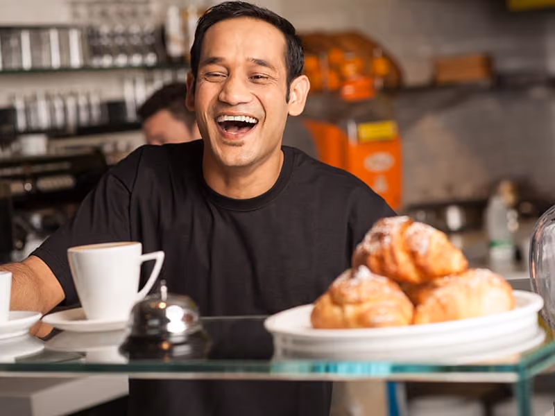 Smiling man behind counter with coffee cup and plate of croissants in a cafe setting.
